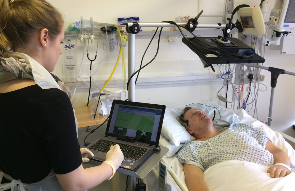 man lying in hospital bed looking at screen, woman with computer by bedside