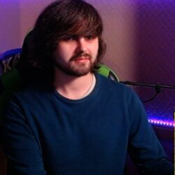 A man in a blue jumper smiles at the camera. He has long brown hair and a beard.