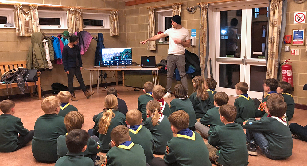 Two standing adults with gaming kit in front of seated group of around 25 children