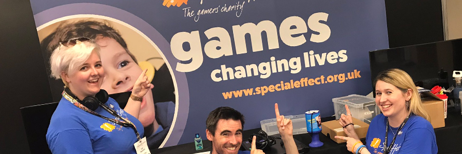 Three smiling people in front of and pointing at an exhibition stand