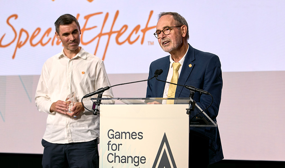 Man standing at a podium on stage giving an award acceptance speech. Another man looks on, holding the award